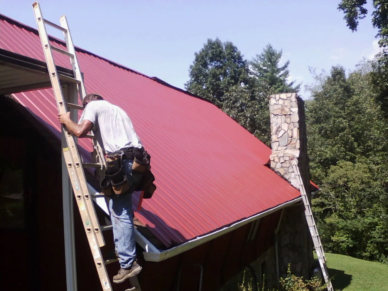 Roofer on a ladder beside a red metal roof and stone chimney — Soffit & Fascia Repair by Roofing Brothers in Rancho Mirage